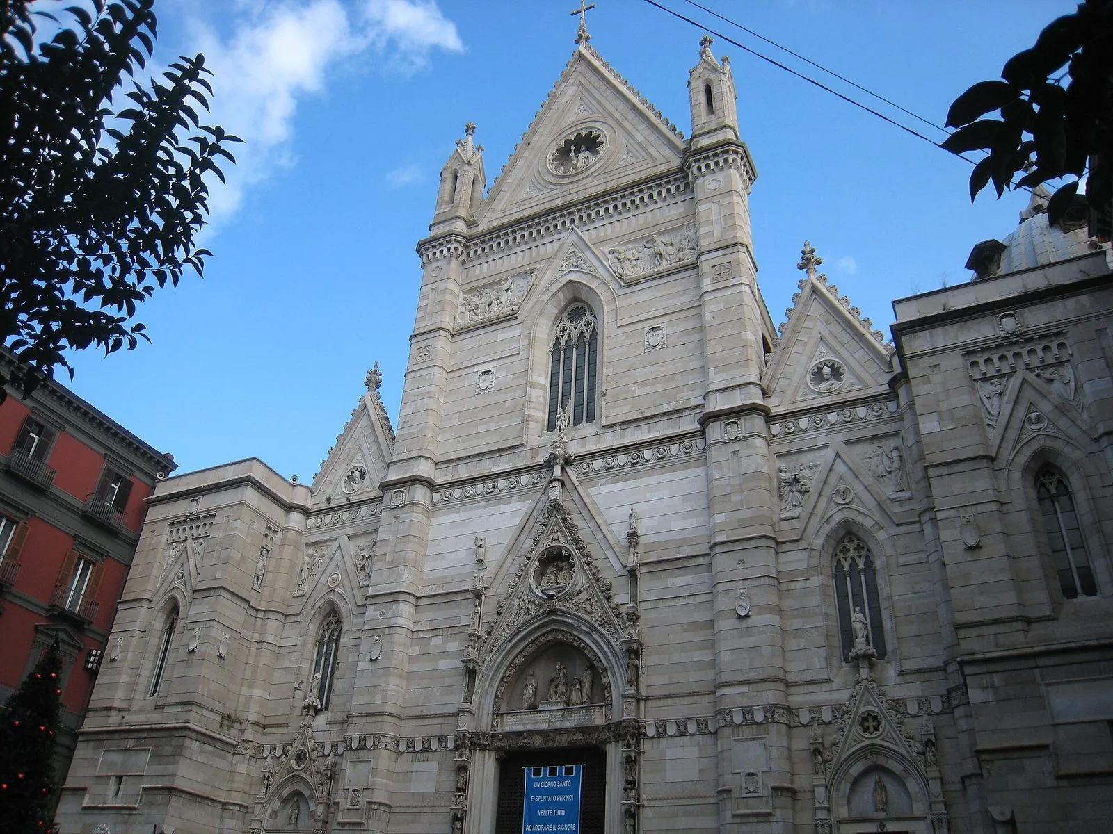 Facciata neogotica in marmo chiaro del Duomo di Napoli vista dal basso, con guglie, rosone e portale centrale, contro un cielo azzurro con nuvole e un palazzo rosso sulla sinistra
