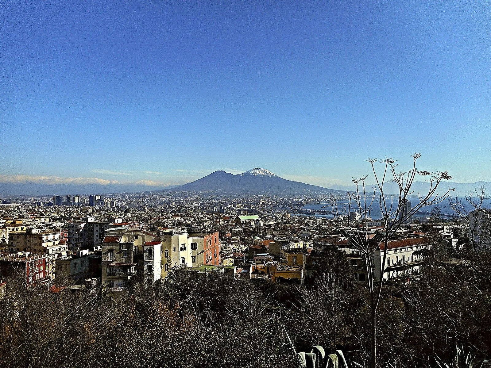 Panorama invernale di Napoli dall'alto sotto un cielo terso: i palazzi della città, il porto e il golfo a destra e, sullo sfondo, il Vesuvio con la cima innevata; in primo piano alberi spogli