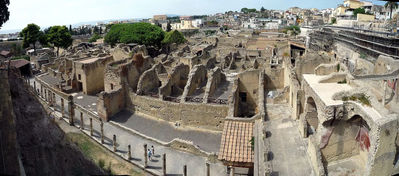 Veduta panoramica dall'alto degli scavi di Ercolano: case romane in tufo senza tetto, un portico di colonne lungo una strada antica, qualche visitatore lontano e la città moderna sul bordo dello scavo
