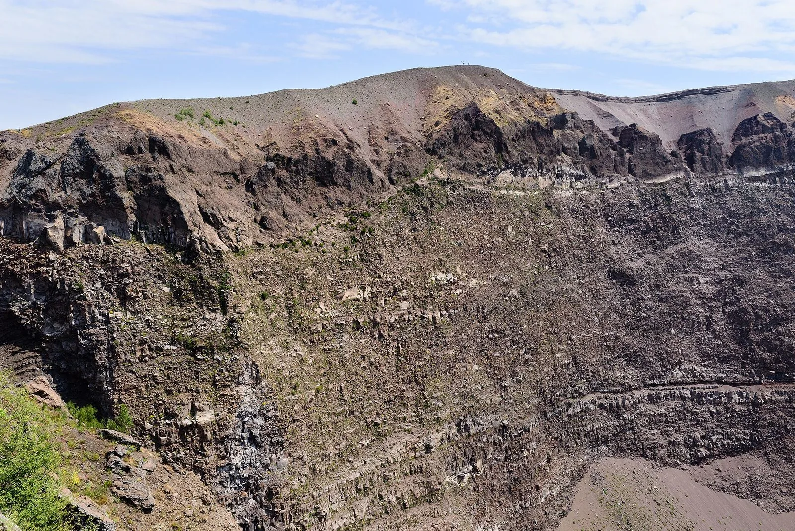 L'interno del cratere del Vesuvio visto dal bordo: pareti ripide di roccia lavica scura e stratificata, ciuffi di vegetazione in primo piano a sinistra e il profilo del cono sotto un cielo azzurro velato